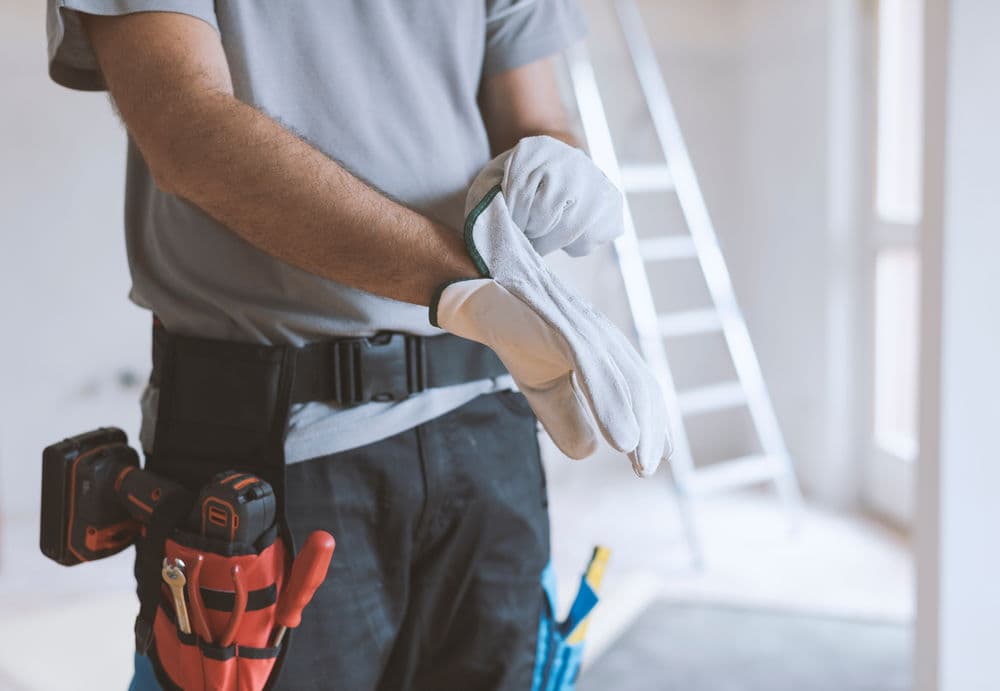 Construction worker wearing gloves, preparing for a project with a tool belt and ladder in background.