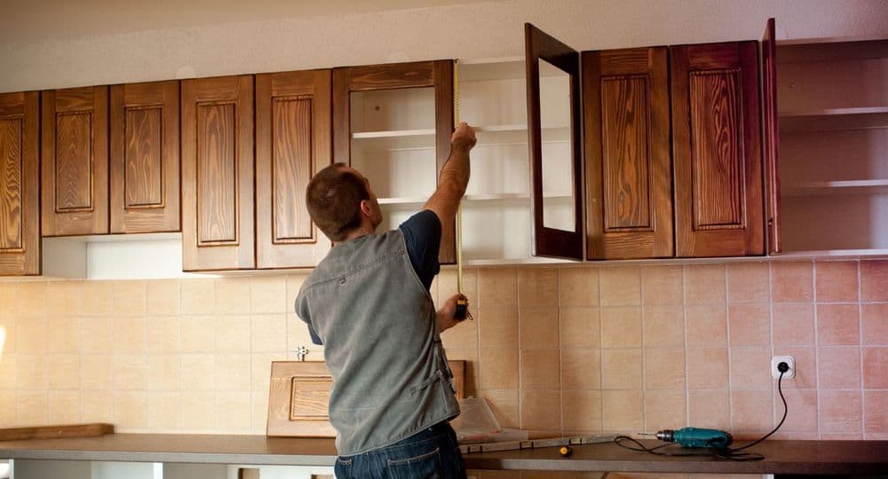 Carpenter installing wooden kitchen cabinets, using a measuring tape and power drill.