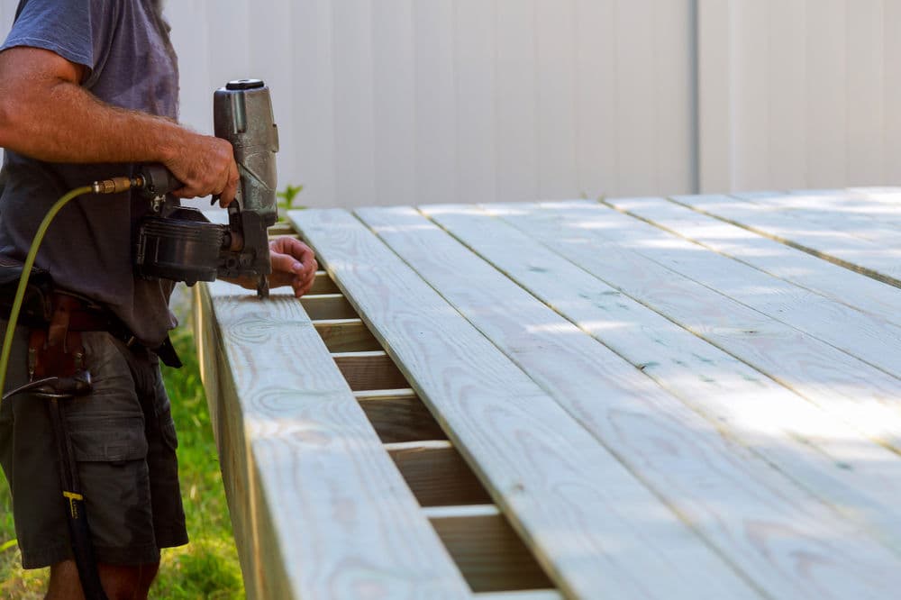 Carpenter using a nail gun to install wooden planks on a deck frame outdoors.